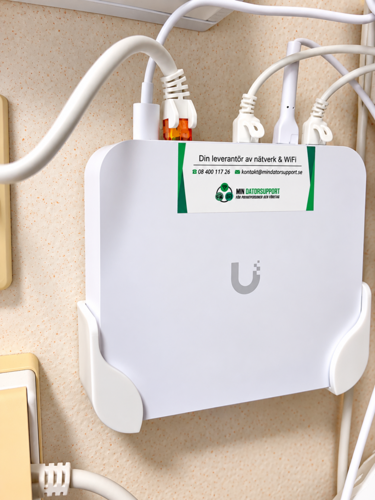 UniFi Gateway Ultra i ett väggfäste bredvid ett gammalt teleuttag, med Min Datorsupports dekal och nätverkskablar